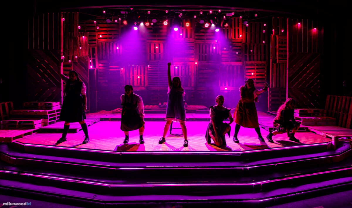 A high, wide view of the ensemble in silhouette on the tiered stage under an intense magenta backlight.