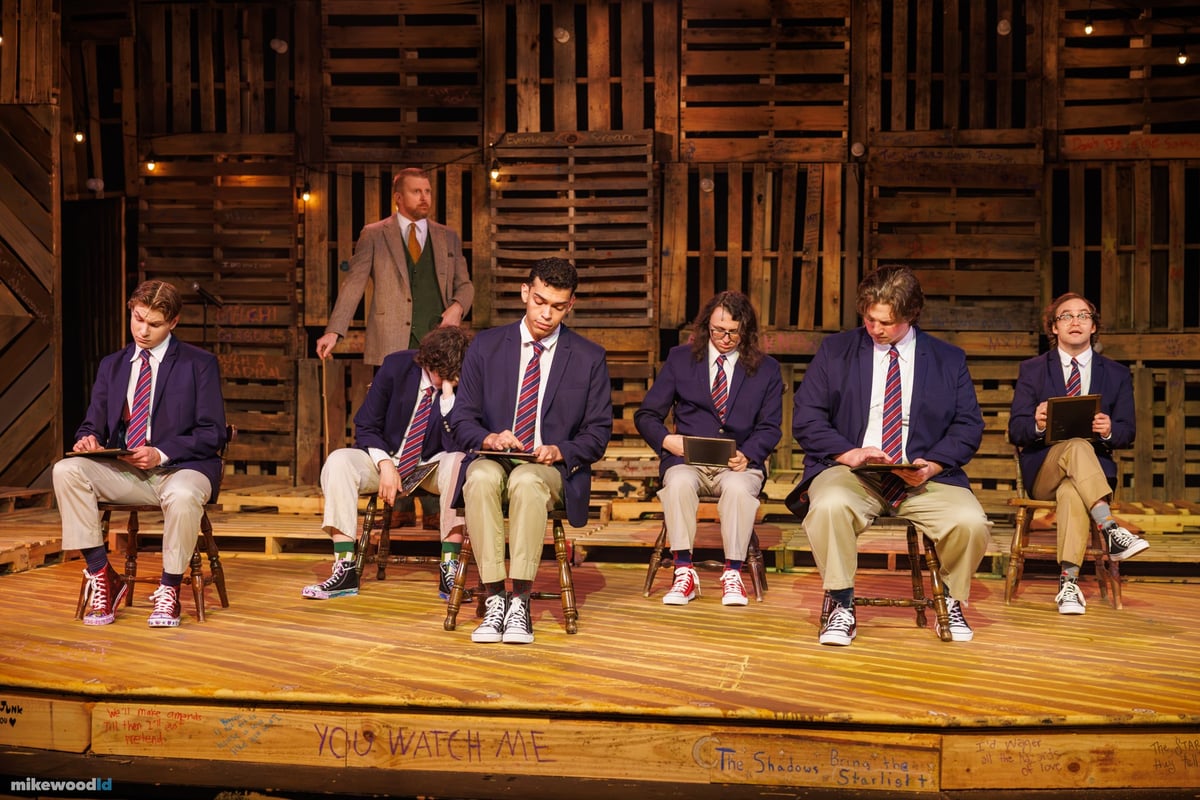 The schoolboys sit in a row in blazers and ties under warm light as a schoolmaster stands behind them at the pallet wall.