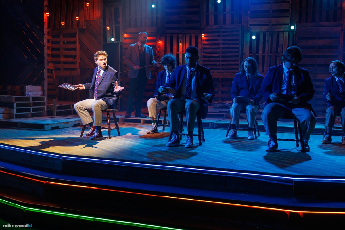 The schoolboys sit across the tiered stage in a cool blue wash edged with green neon striping, one boy forward on a stool with a book.