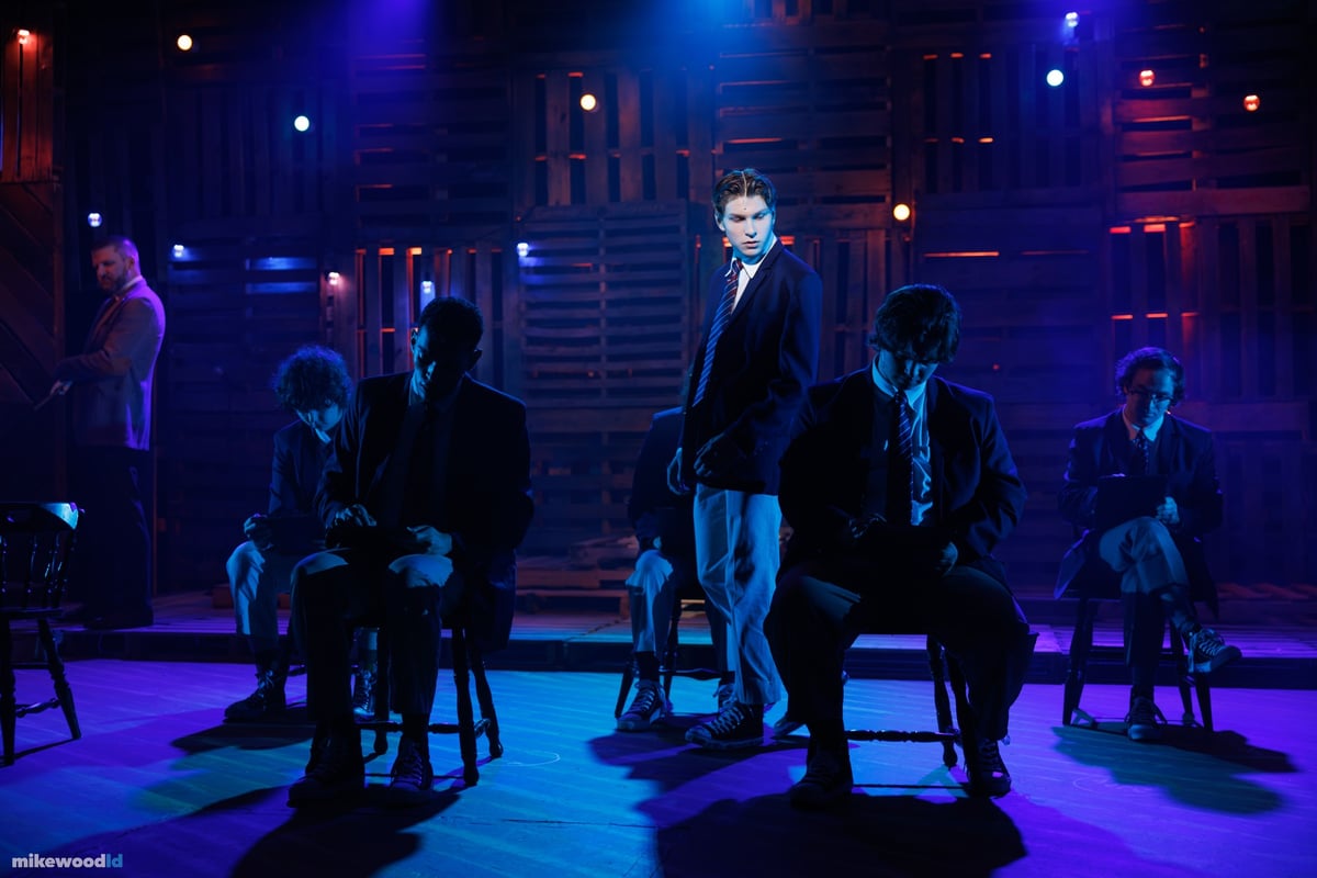 The boys of the company fill the stage in deep blue and teal light, a single figure spotlit standing among them.