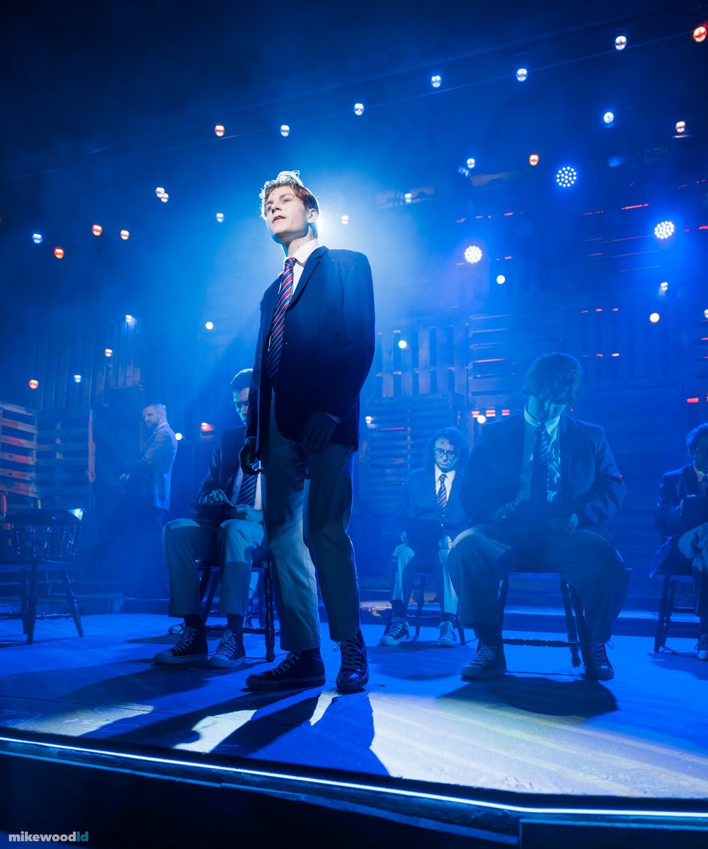 A young man stands alone in a hard shaft of blue light, hands in his pockets, the rest of the cast dim behind him.