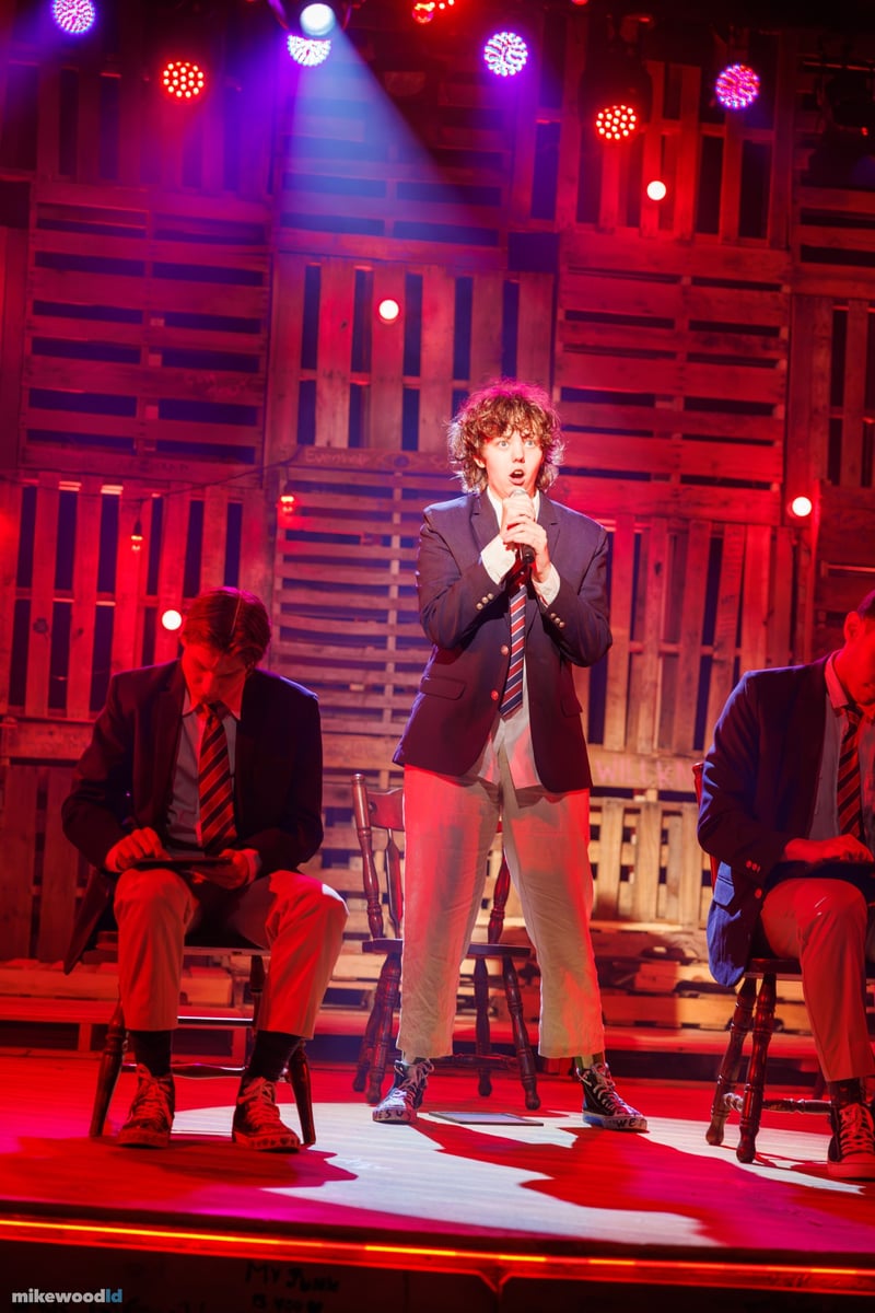 A curly-haired young man in a blazer sings into a handheld microphone against a red-washed pallet wall.