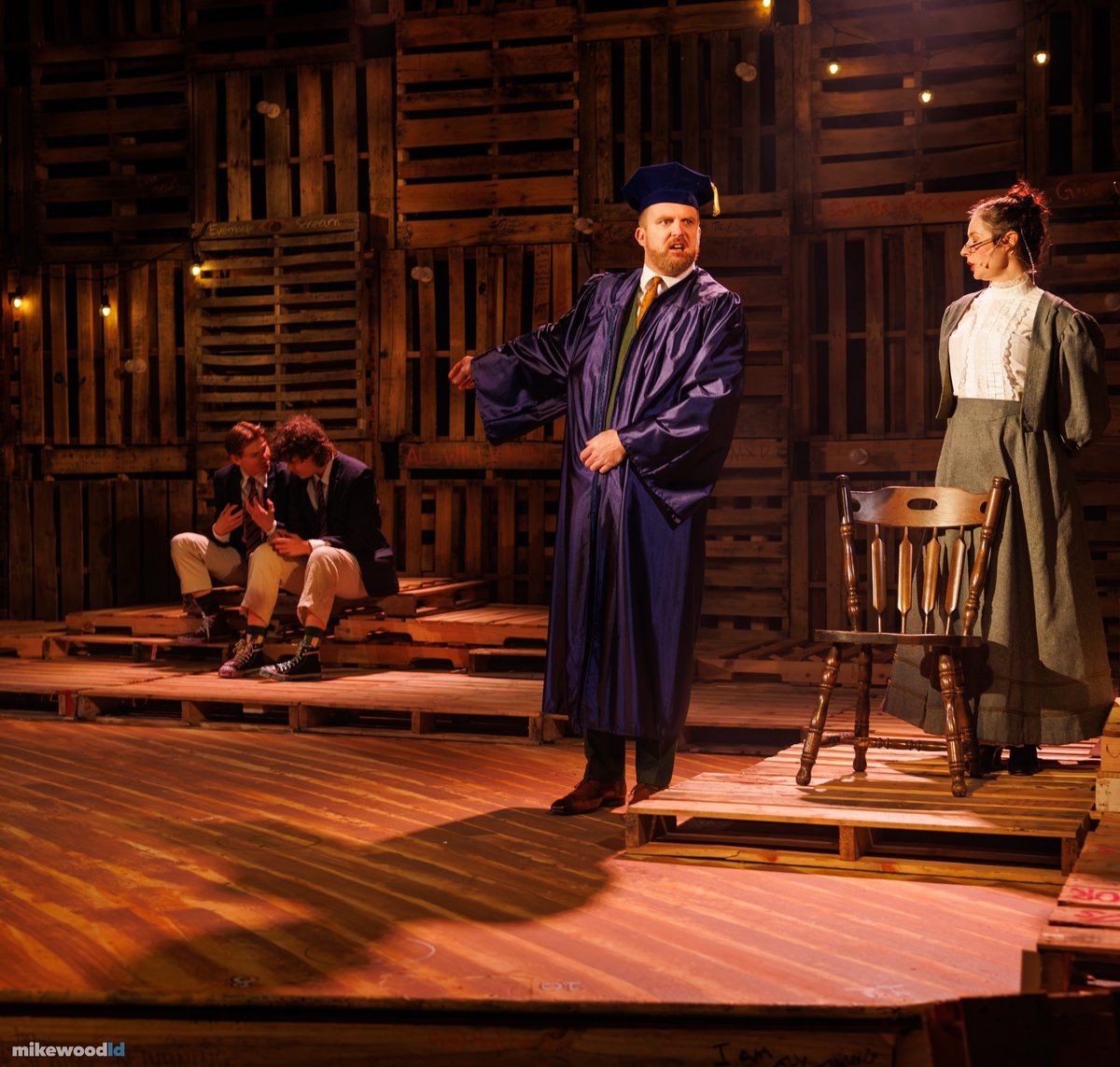 A figure in an academic gown and mortarboard presides over students seated on the floor in warm light.