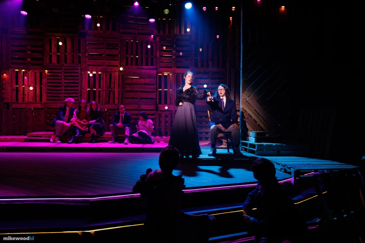 A lone spotlit figure stands on a platform amid teal and violet haze on an otherwise dark stage.