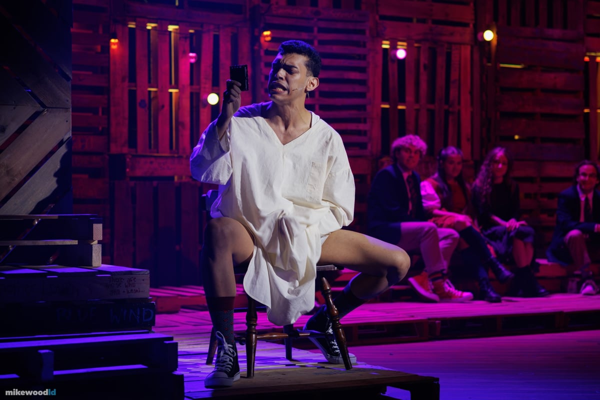 A young man in a white nightshirt kneels on a chair holding up a light, ringed by a magenta and purple wash.