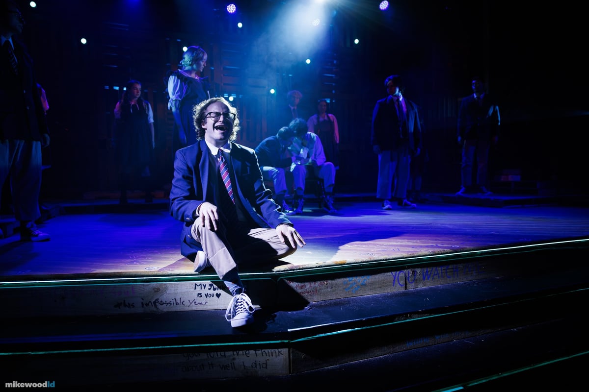 A young man sits slumped and alone on the plank steps in a tight spotlight, the cast dim in the darkness behind.