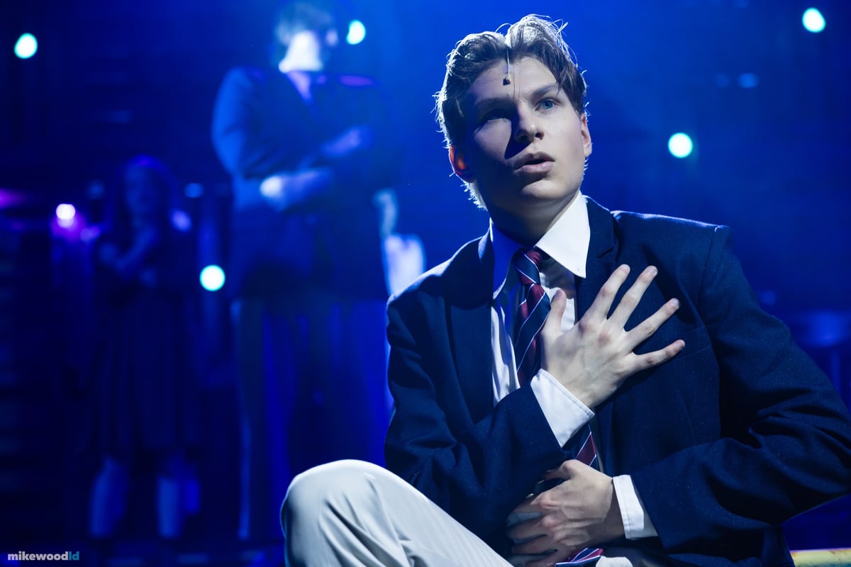 A tight close-up of a young man kneeling with a hand pressed to his chest, lit in cool blue.