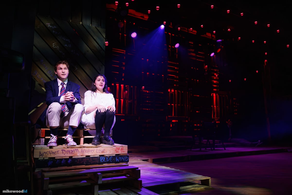 A young man and young woman sit close together on a low pallet platform in a soft purple wash.