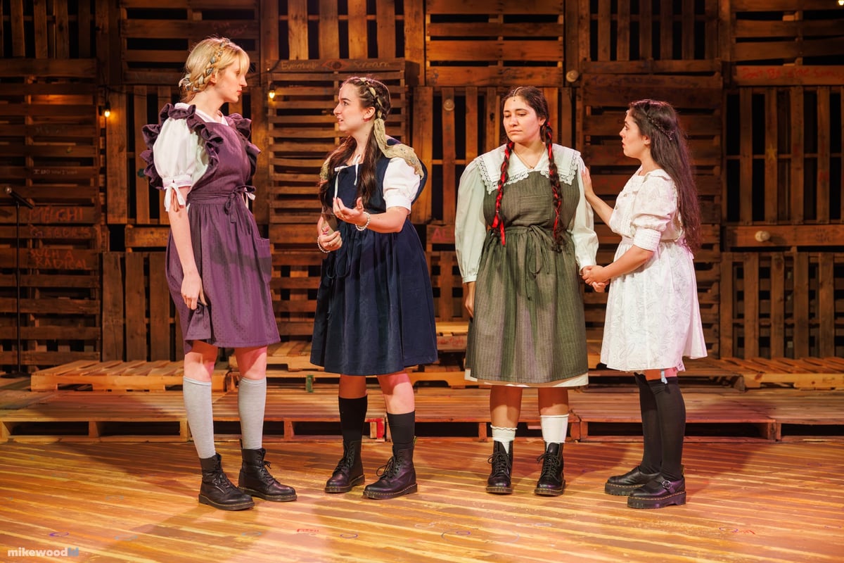 Four young women in period school dresses stand in a line holding hands under warm amber light.