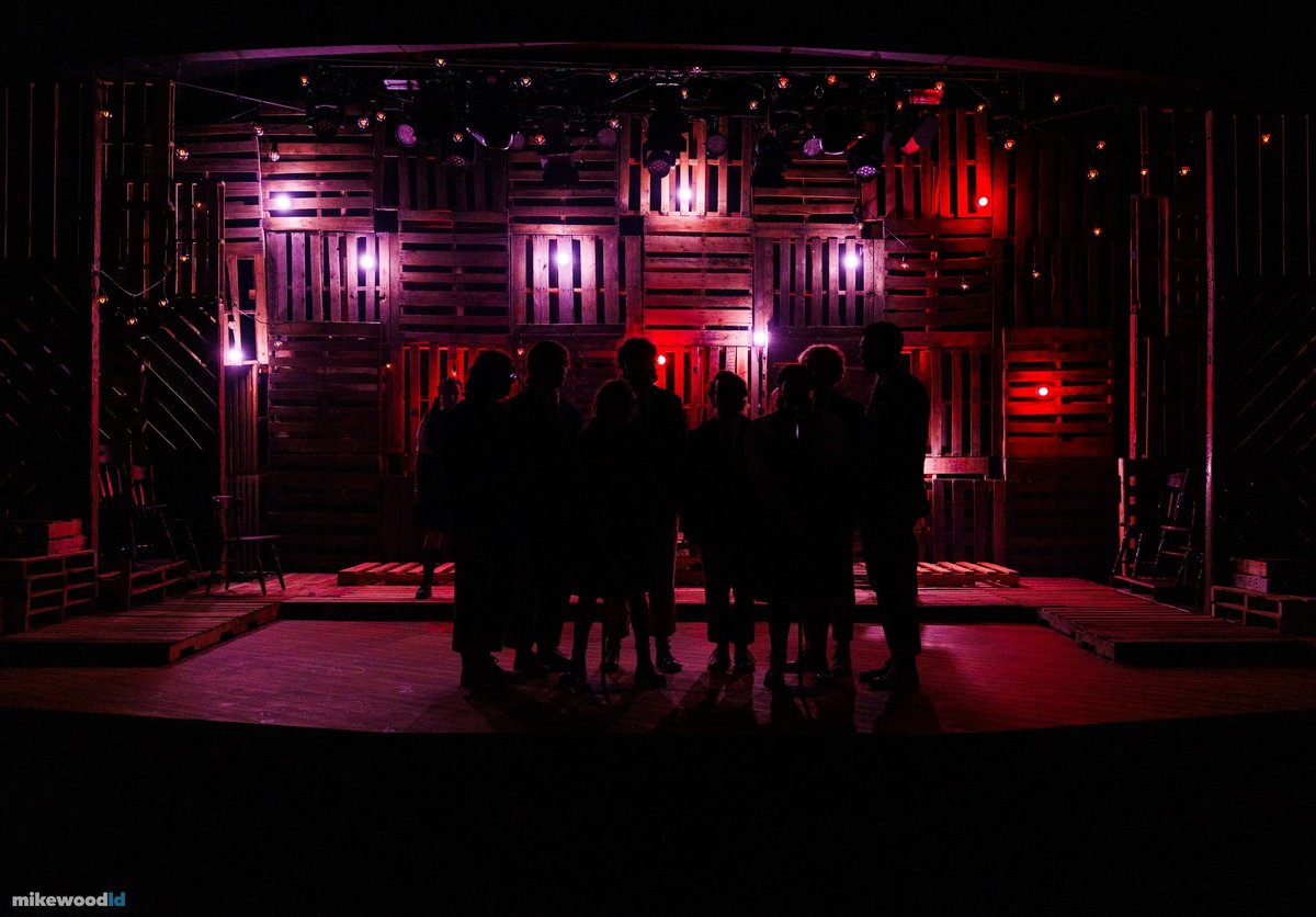 The company stands clustered in silhouette facing a red-lit pallet wall in low, dramatic light.