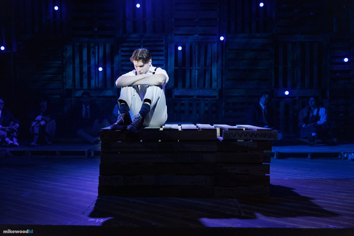 A young man sits hunched atop a pallet cube, knees drawn up, isolated in a pool of blue light.
