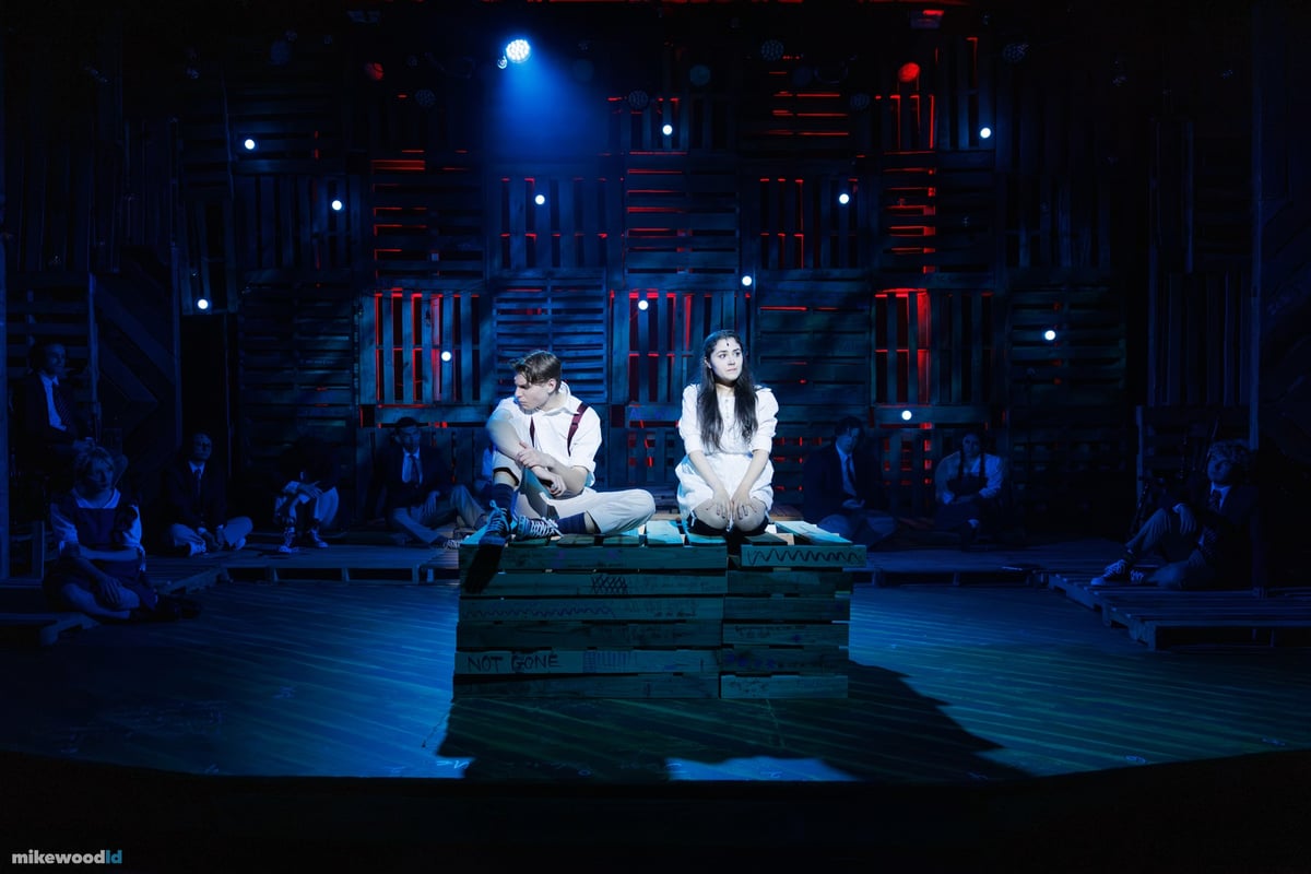A young man and young woman sit together on a pallet cube under a blue spotlight, the ensemble seated in the surrounding dark.