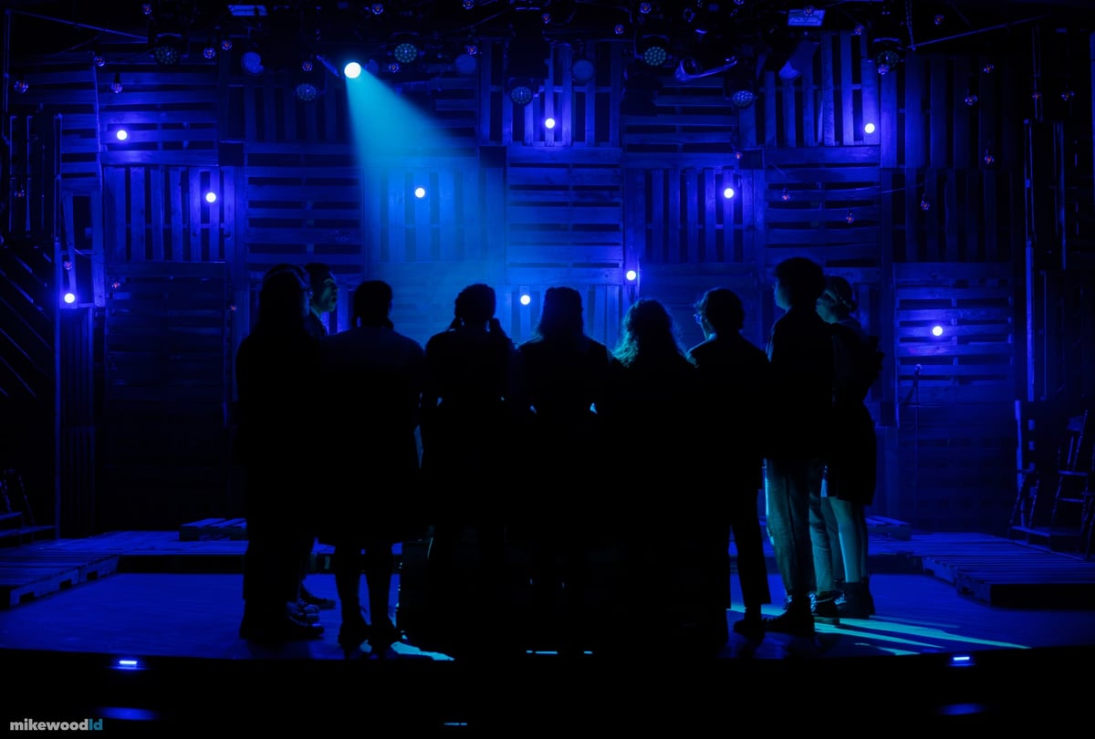 The company stands in silhouette amid blue beams and haze on a darkened stage.