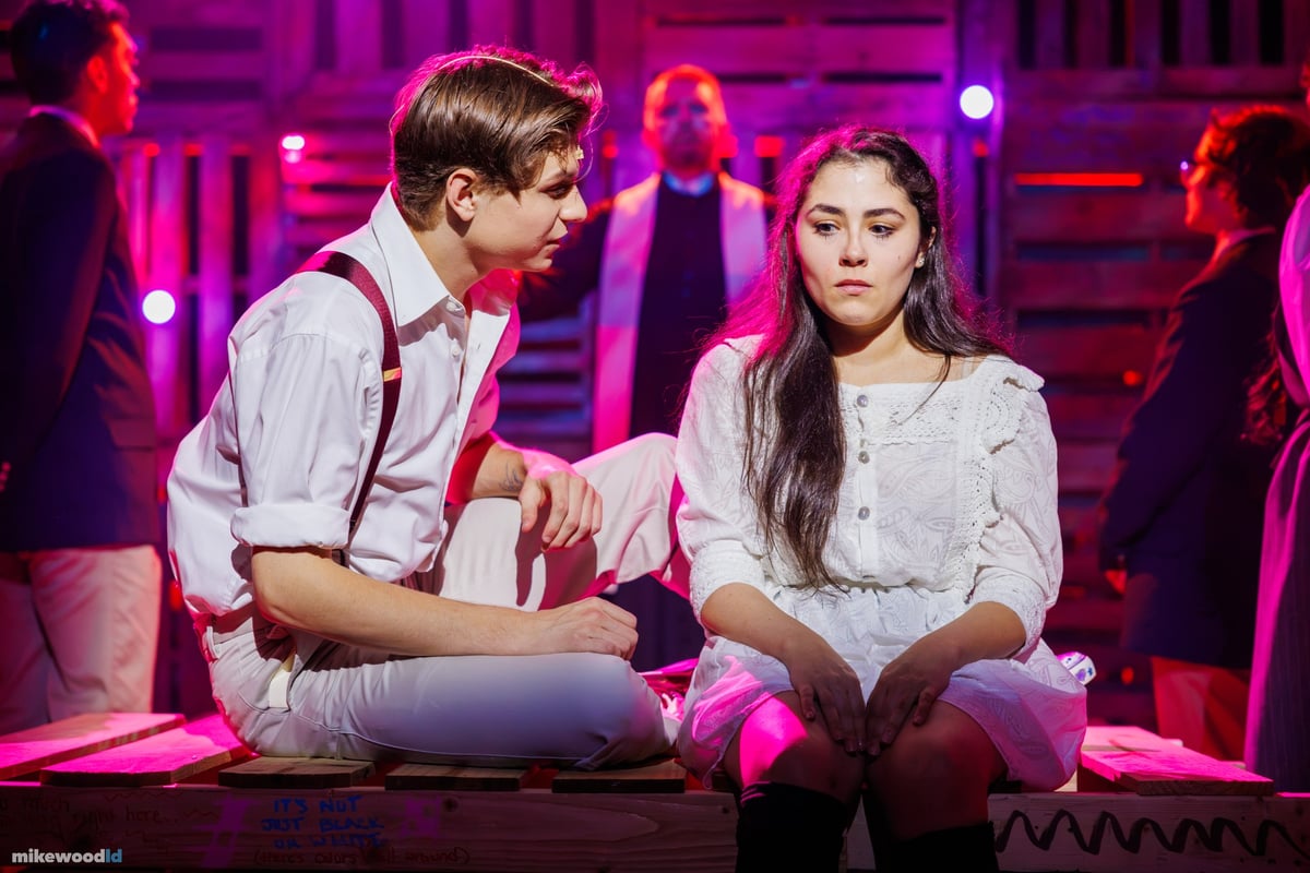 A young man in suspenders sits cross-legged beside a young woman in white amid a magenta wash, the cast around them.