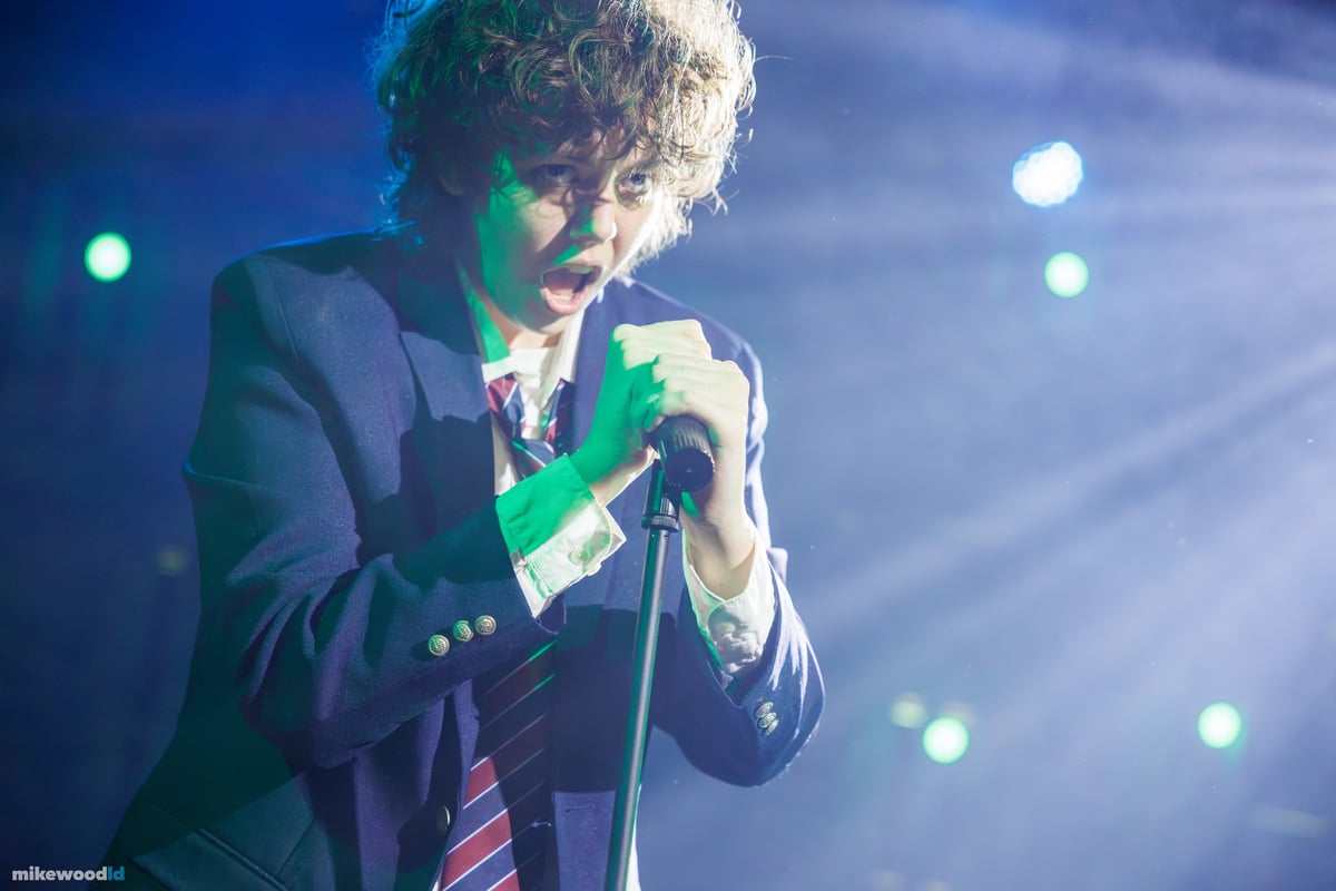 A close-up of a curly-haired young man singing into a handheld microphone in a green-white beam and haze.
