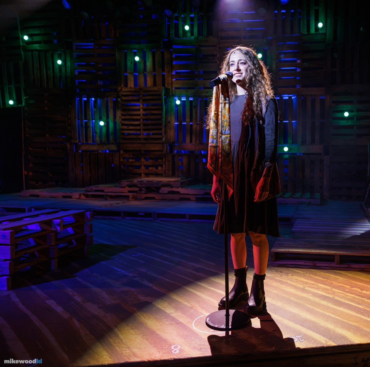 A young woman in a dark coat sings at a microphone in a tight downlight, green and blue accents on the pallet wall behind.