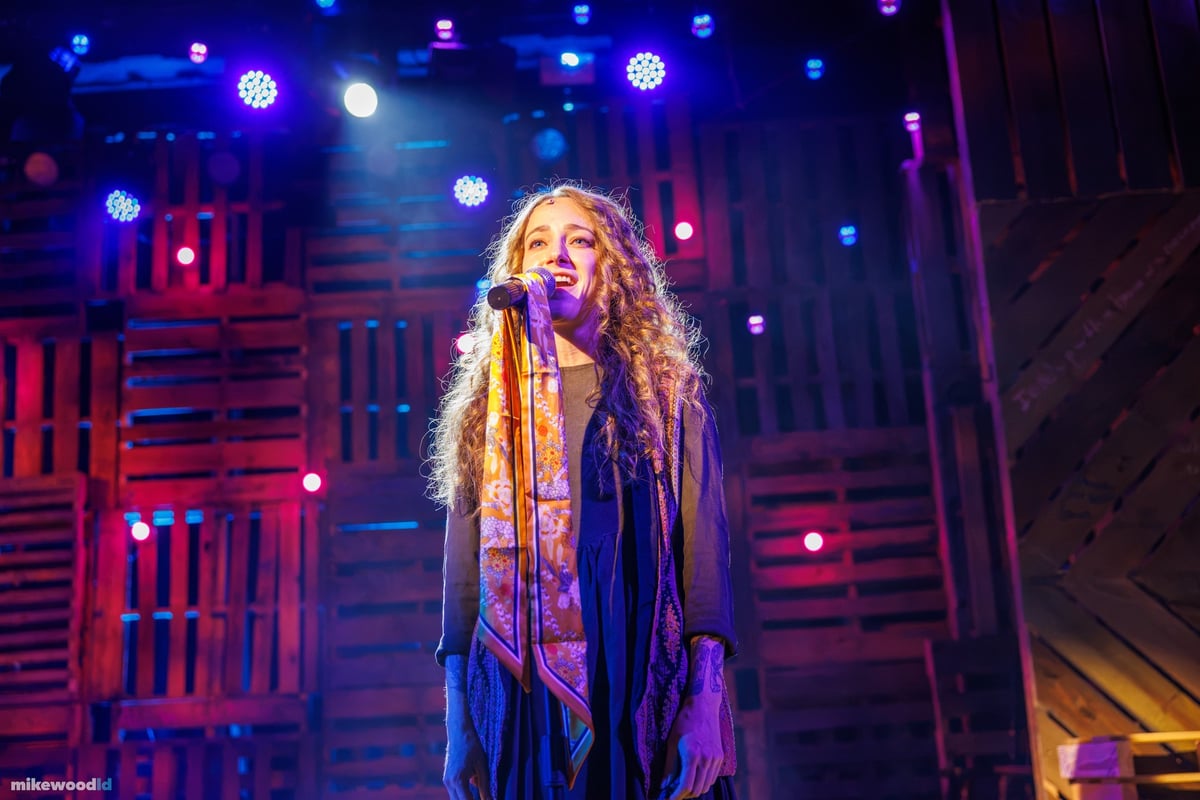 A young woman with long curly hair sings at a microphone against a pink and purple pallet-wall wash.