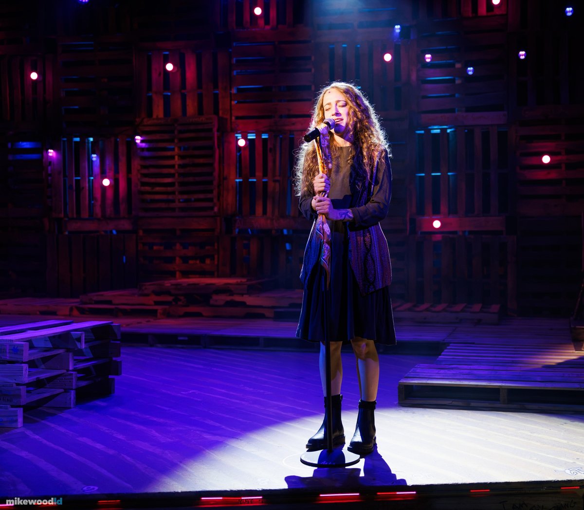 A young woman sings at a microphone in a narrow downlight on the open stage, the pallet wall dim behind.