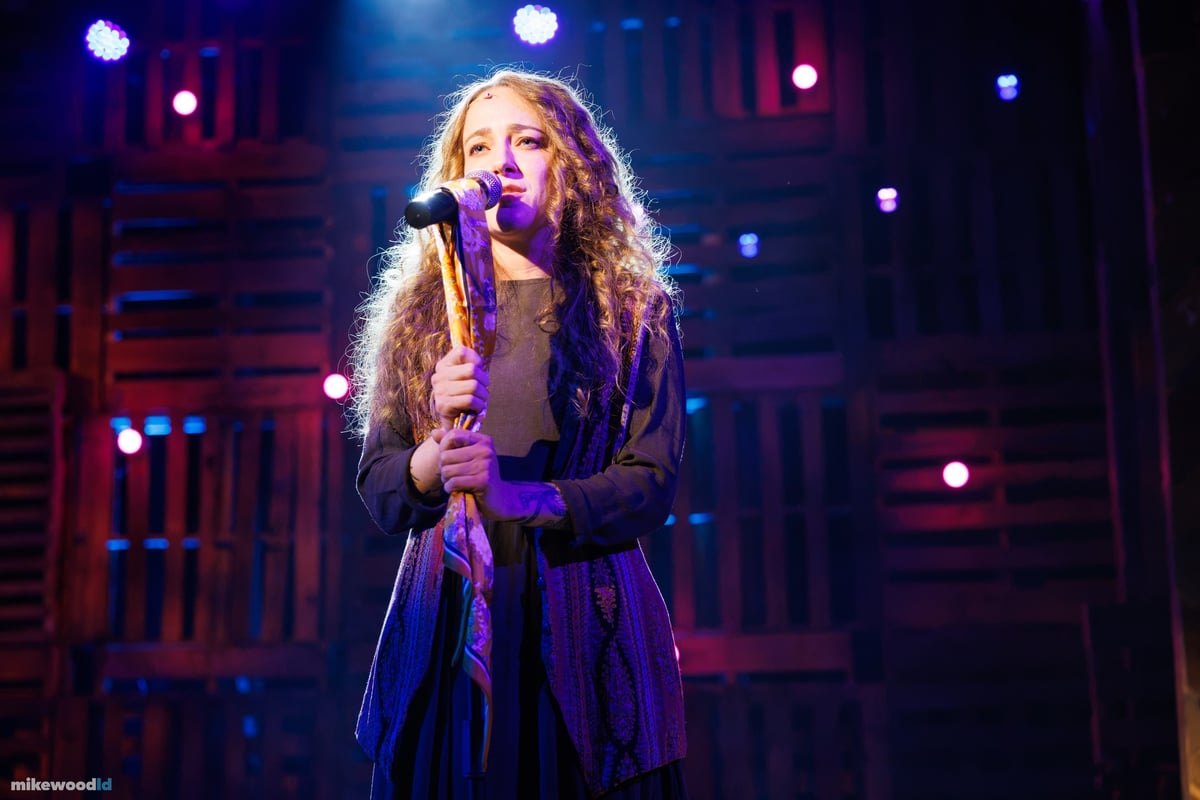 A close-up of a young woman singing into a handheld microphone in a purple and blue wash.