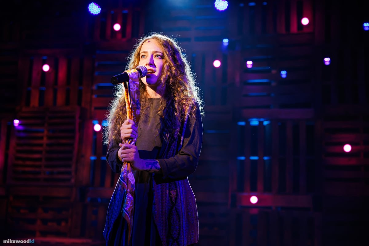 A young woman sings into a handheld microphone, lit in blue and violet against the pallet wall.