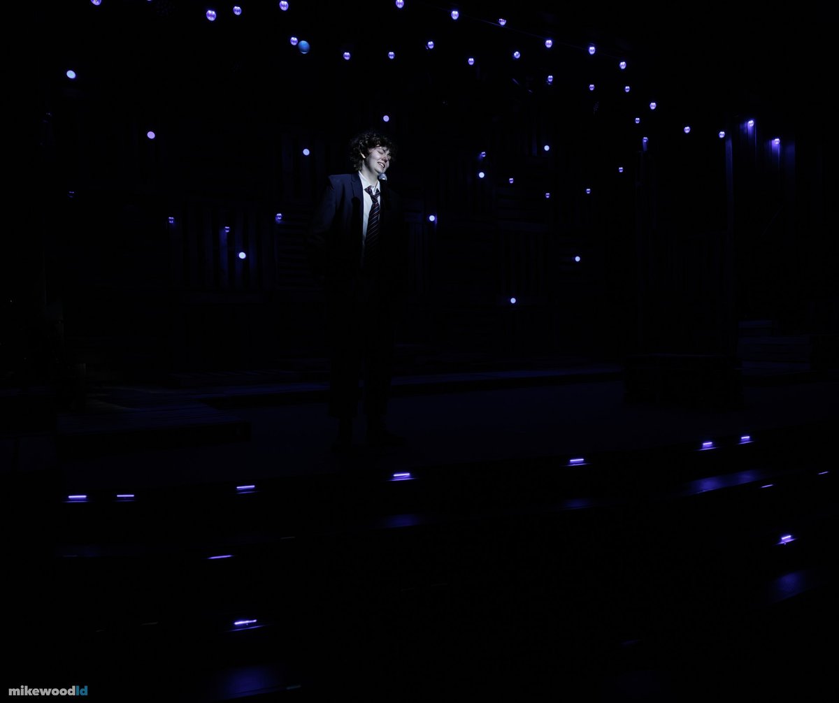A single small figure stands spotlit far upstage on a vast dark stage scattered with star-like pinlights.