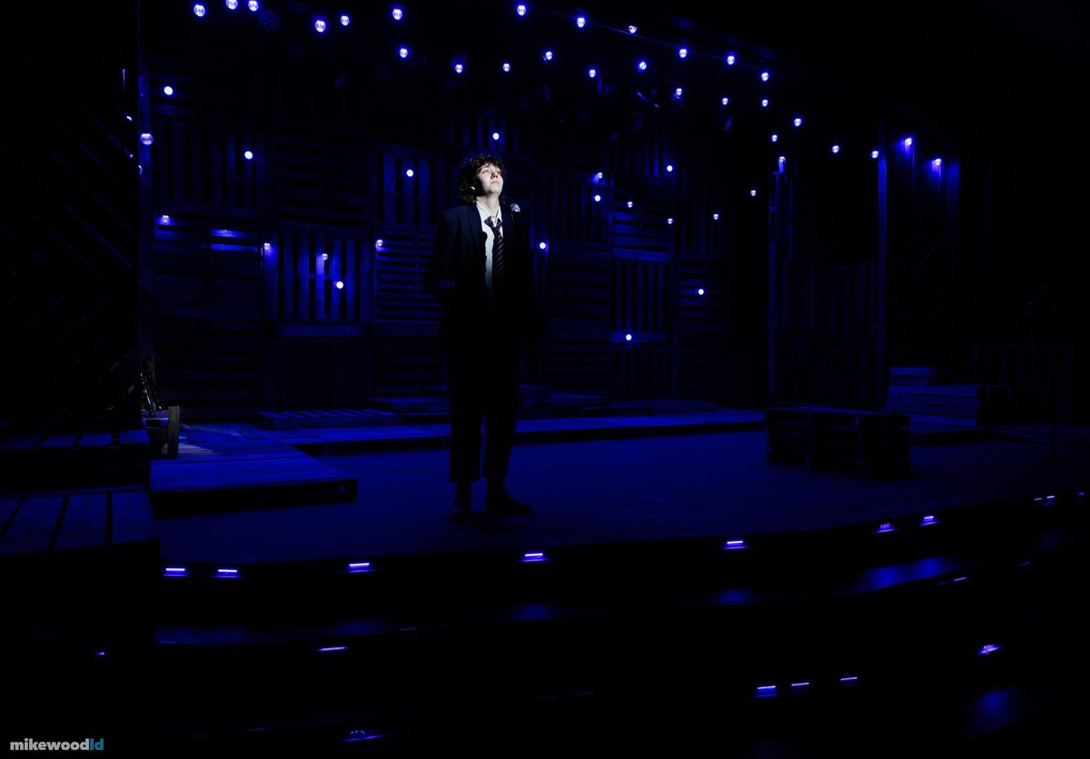 A lone figure stands spotlit at center in the darkness, surrounded by pinlight stars.