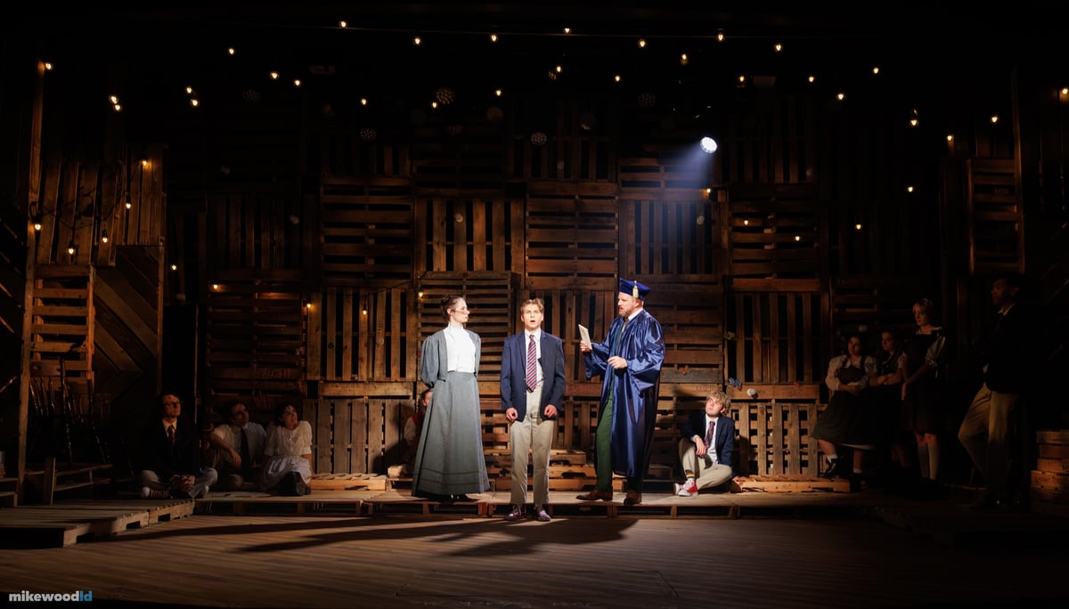 A confrontation plays out center stage in warm light, a gowned figure among them, as the cast watches from the dark.