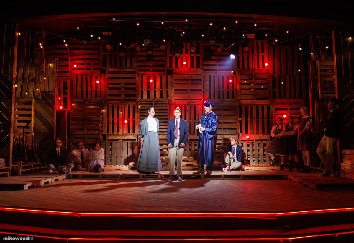 A group scene unfolds center stage against a red-washed pallet wall, the ensemble seated around the perimeter.