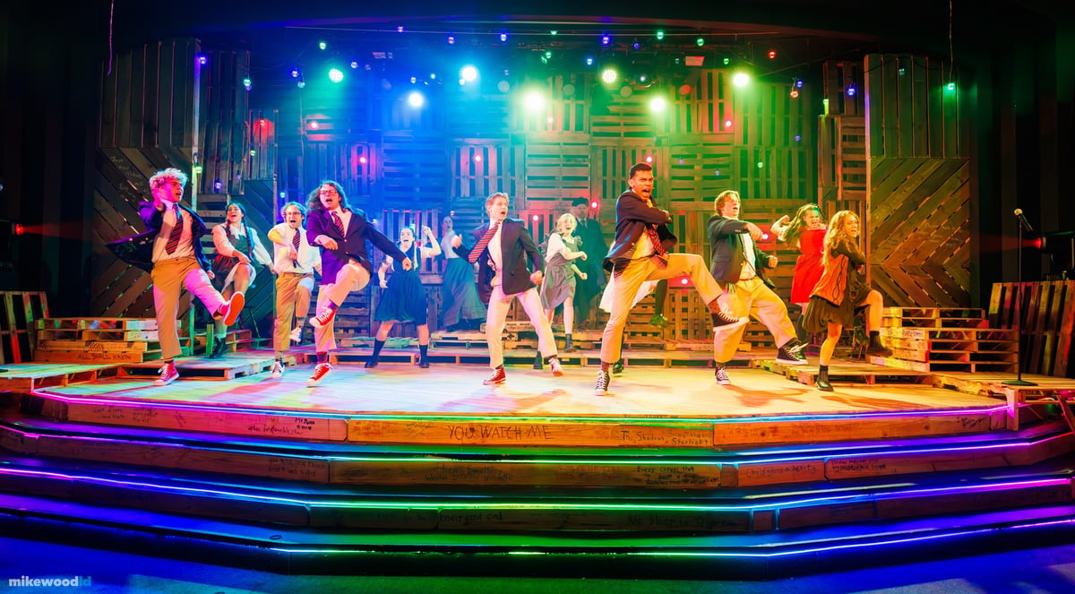 The full company leaps in unison across the tiered stage under a vivid rainbow wash.