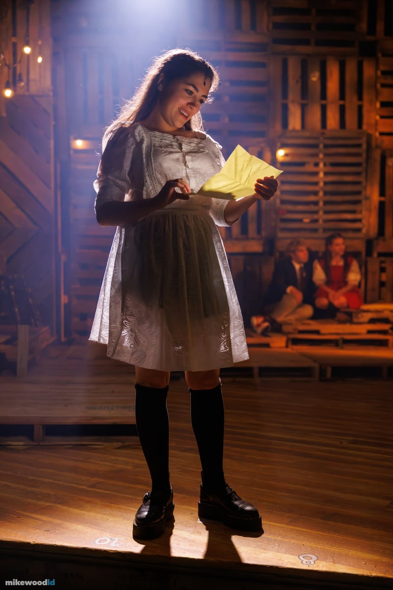 A young woman in a period dress reads a letter in a warm downlight, the cast dim behind her.