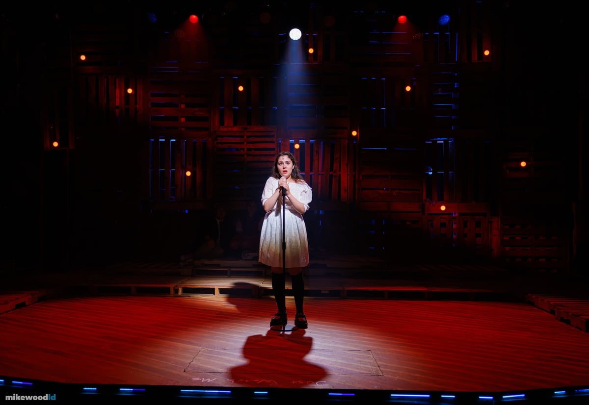 A young woman sings at a microphone in a tight downlight on a wide, dark stage rimmed in blue.