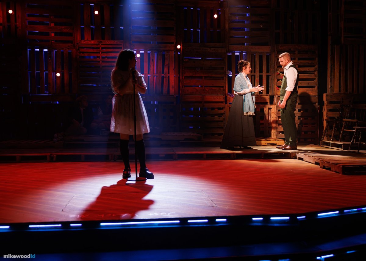 A young woman sings at a microphone in a pool of red light as two figures talk upstage.
