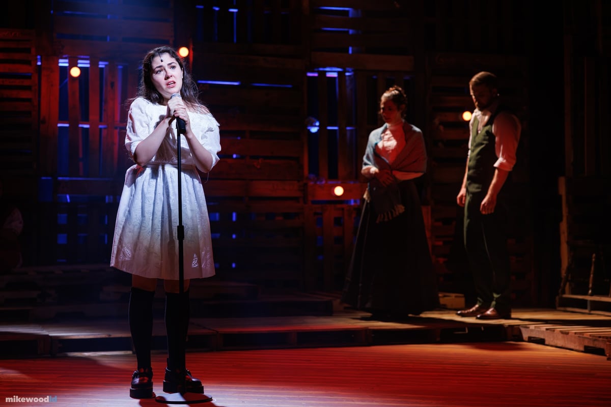 A young woman in a white dress sings at a microphone in red and blue light, two figures behind her.