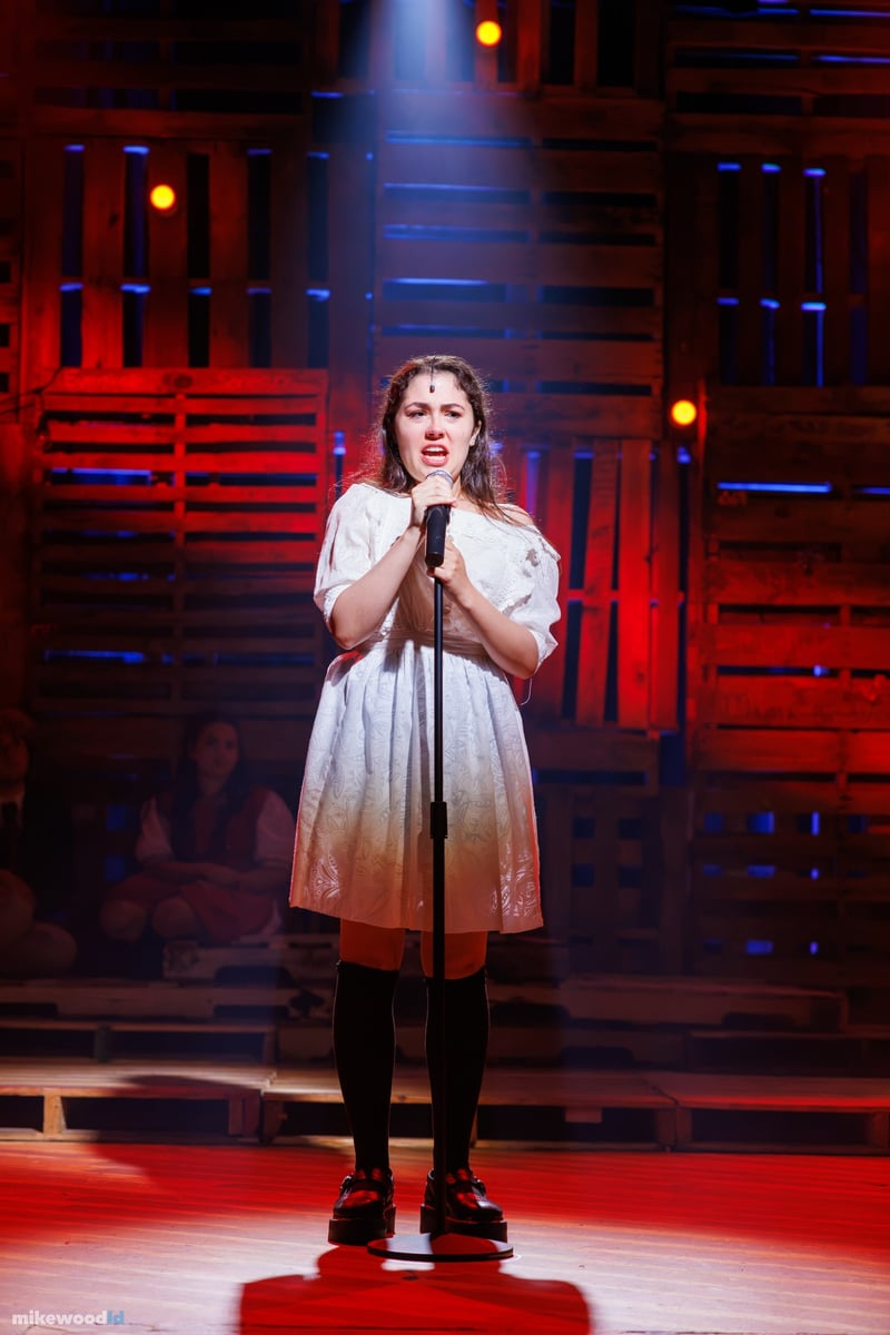 A young woman in a white dress stands at a microphone with hands clasped, lit in red and blue.