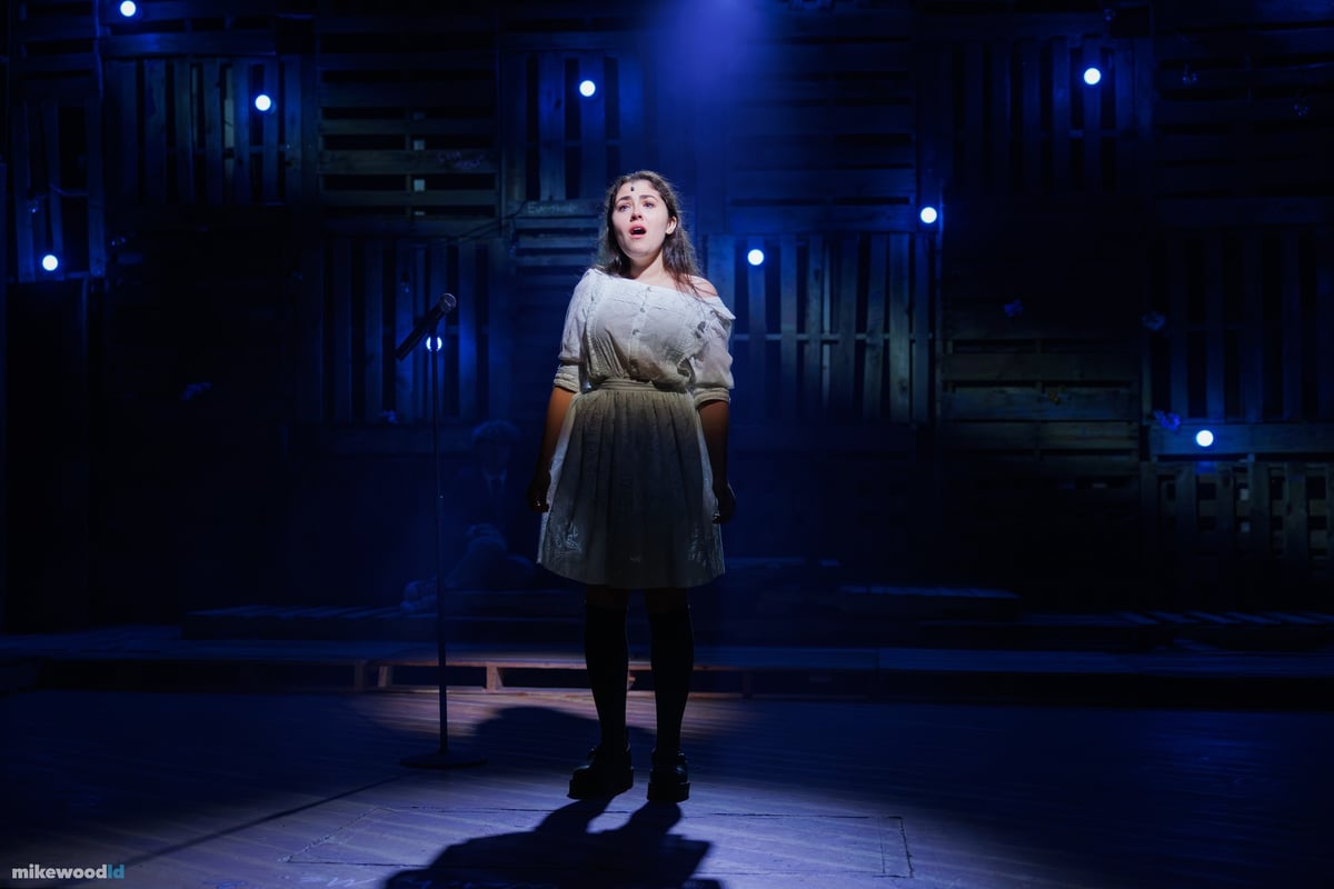 A young woman in a period dress sings alone in a blue spotlight amid star-like pinlights on the dark stage.