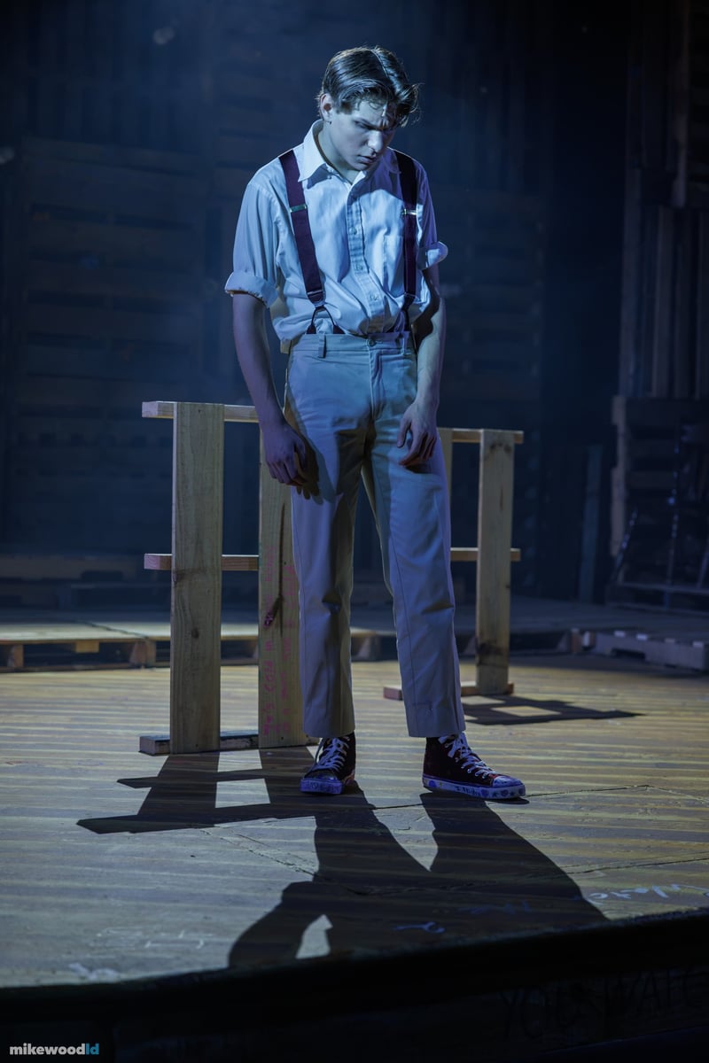 A young man in suspenders stands over a wooden stool in cool blue-grey light, casting a long shadow.