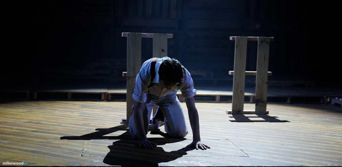 A young man crouches to the floor among wooden frames in a tight spotlight on the dark stage.