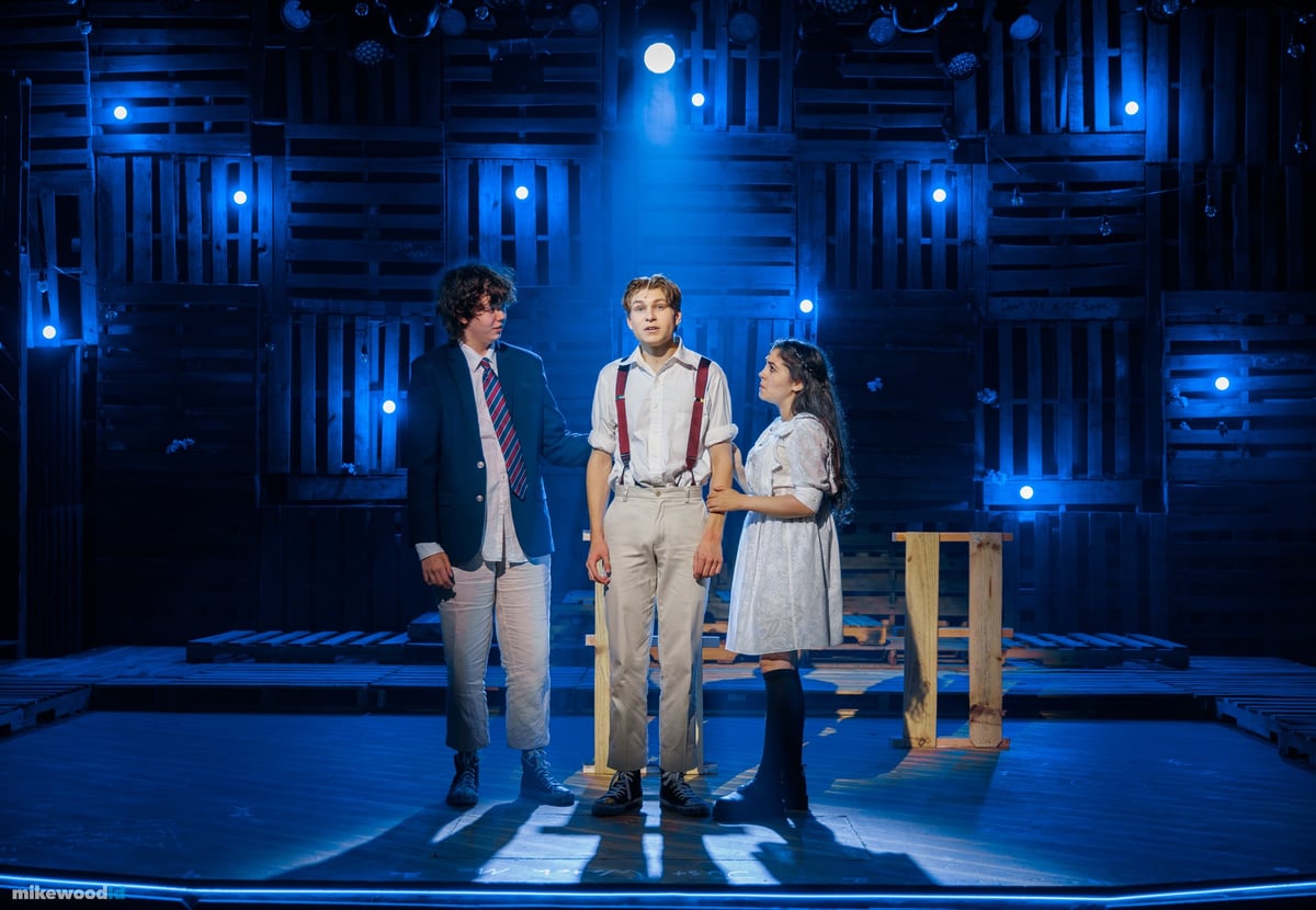 Three young performers stand together center stage in a blue wash, the pallet wall glinting with blue pinlights behind them.