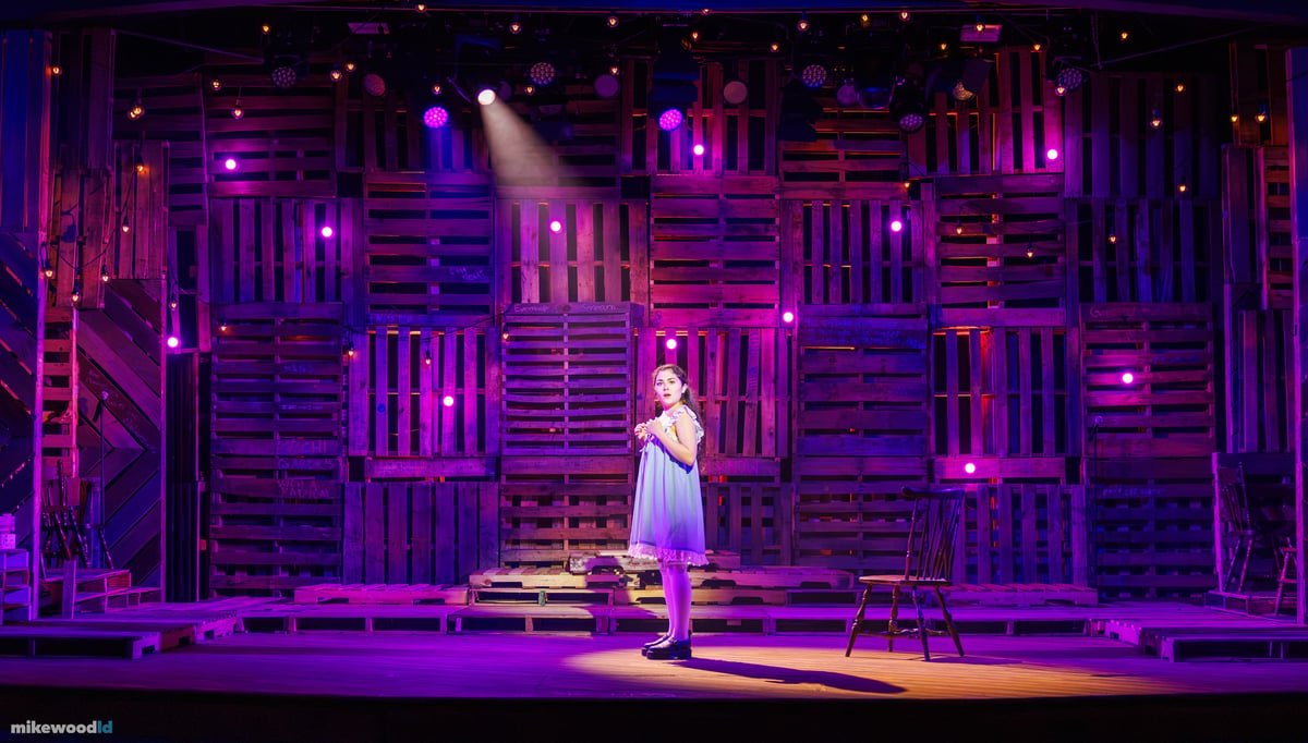 A young woman in a pale blue dress stands alone in a tight spotlight at center stage, the pallet-wood wall behind her dotted with warm practical bulbs under a magenta wash.