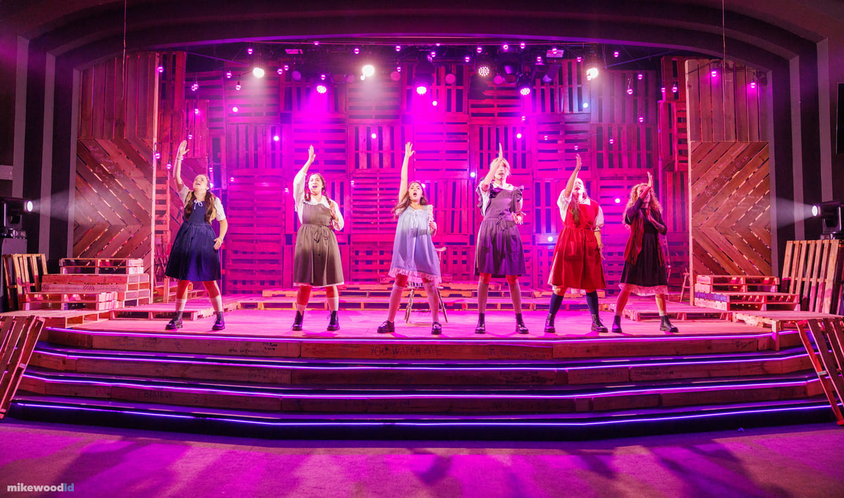 Six young women in period school dresses stand in a line with arms raised, lit in a hot magenta wash as blue and magenta LED striping glows along the step edges.