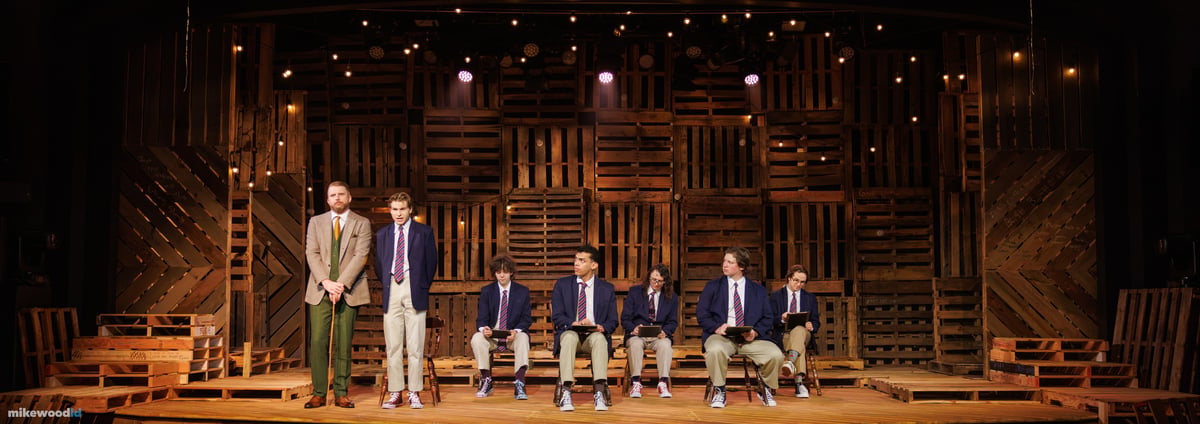 In warm amber light, a schoolmaster in a tweed suit stands at left with a cane as a row of schoolboys in blazers sit on wooden chairs holding slates against the pallet-wood wall.
