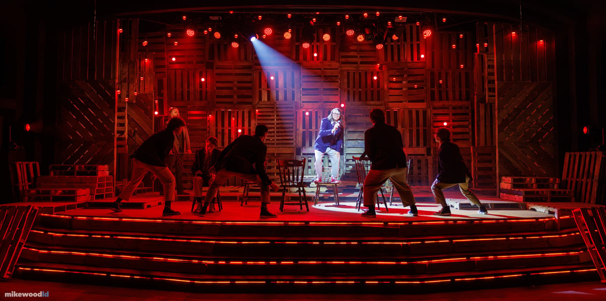 The boys crouch in a ring around a single figure spotlit atop a chair at center, the whole stage saturated deep red with red practicals and red LED striping along the steps.