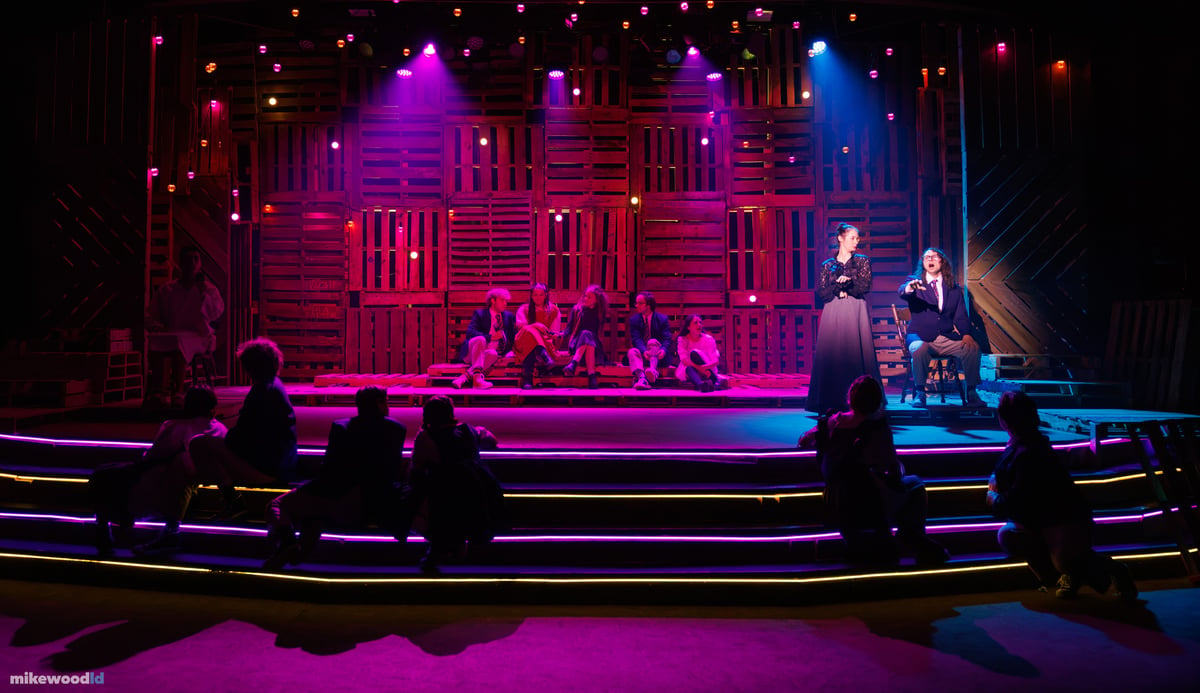 A young woman in a long dark dress stands beside a seated young man at stage right as the ensemble watches from the neon-edged steps, all under a magenta and blue wash.