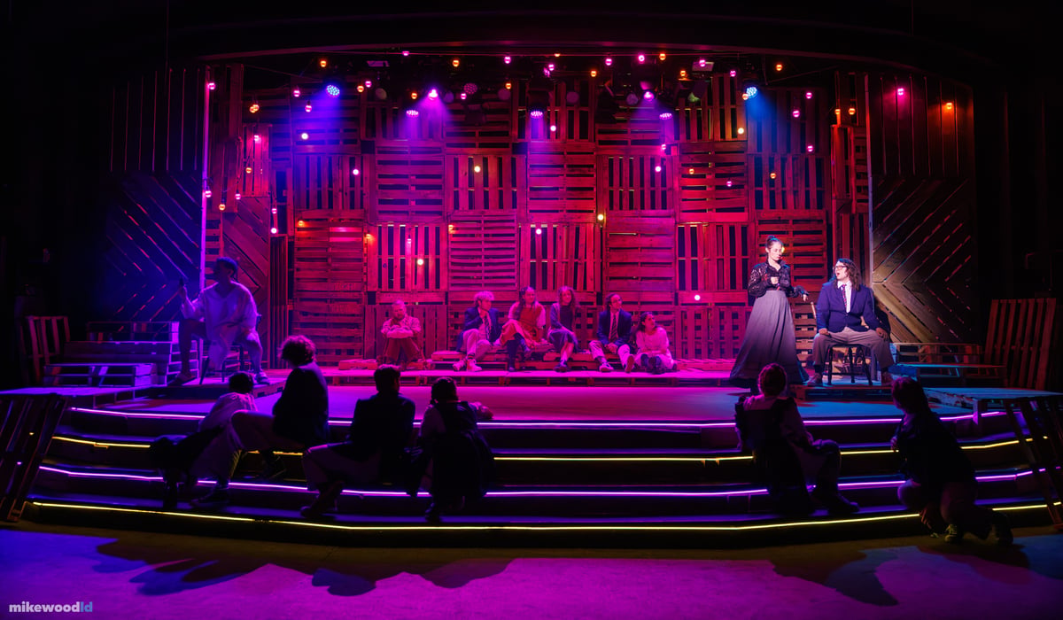 A scene plays out at stage right as the ensemble sits scattered across the pallet platform and neon-edged steps in a deep magenta and blue wash.