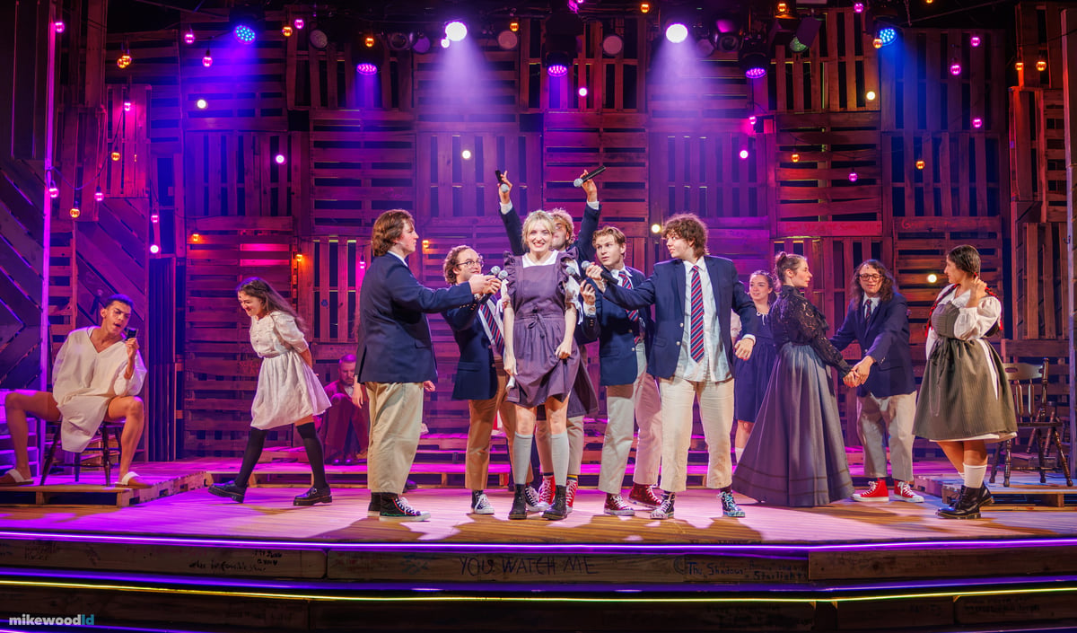 The full company clusters at center with microphones raised under a purple and magenta wash, one figure seated apart on a chair at far left.