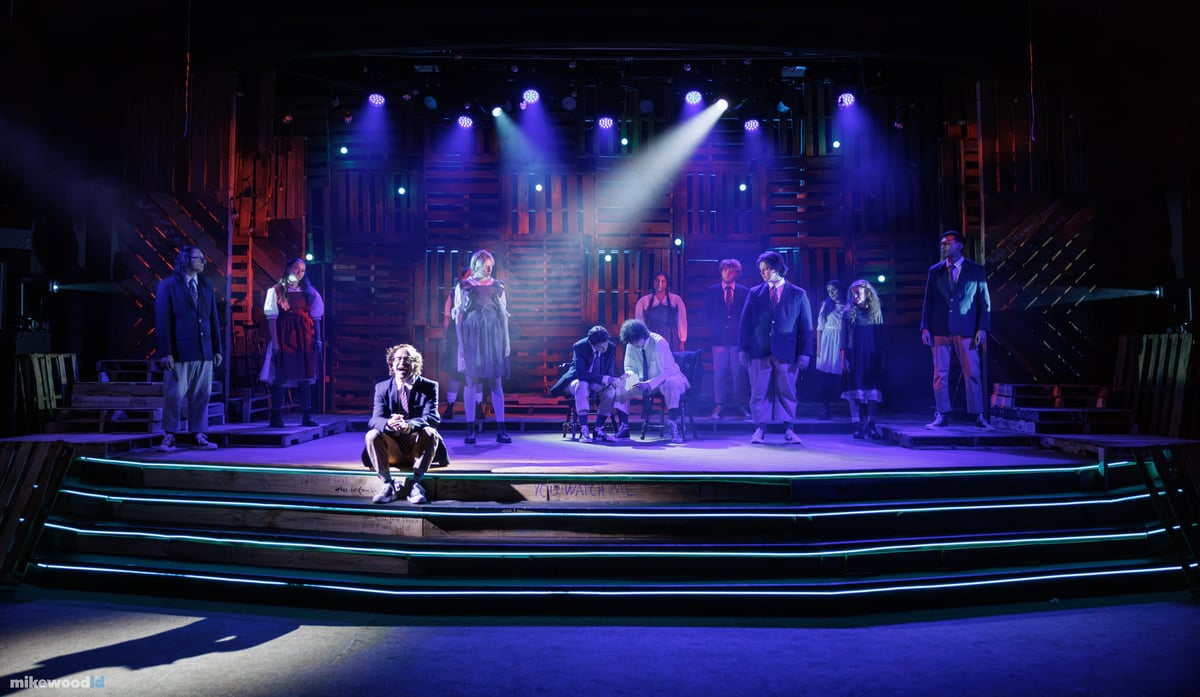 A young man sits alone on the LED-striped steps in a tight top special as the ensemble stands scattered across the stage under a cool blue and violet wash.