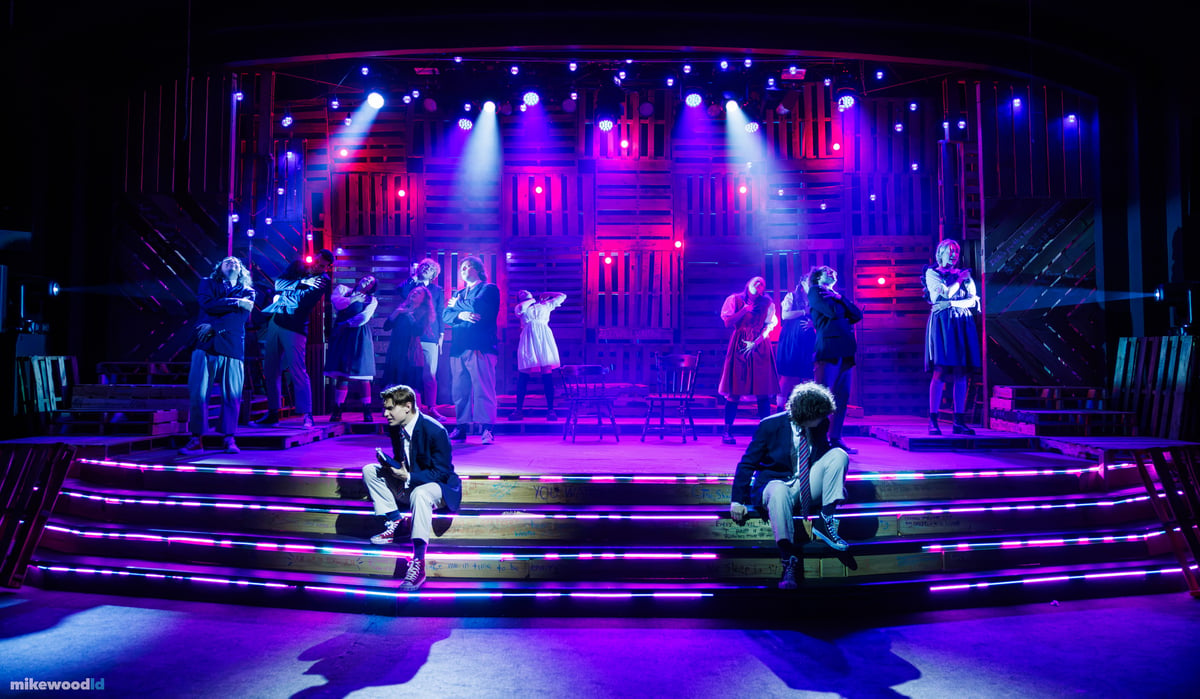 Two young men sit apart on the brightly LED-striped steps as the ensemble stands paired across the stage behind them under a blue and magenta wash.