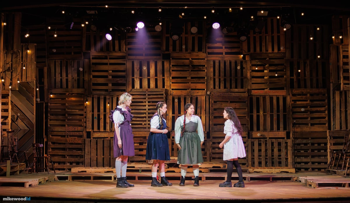 Four young women in period dresses stand in a line in conversation under a soft amber top light, the pallet wall glowing with warm practical bulbs.