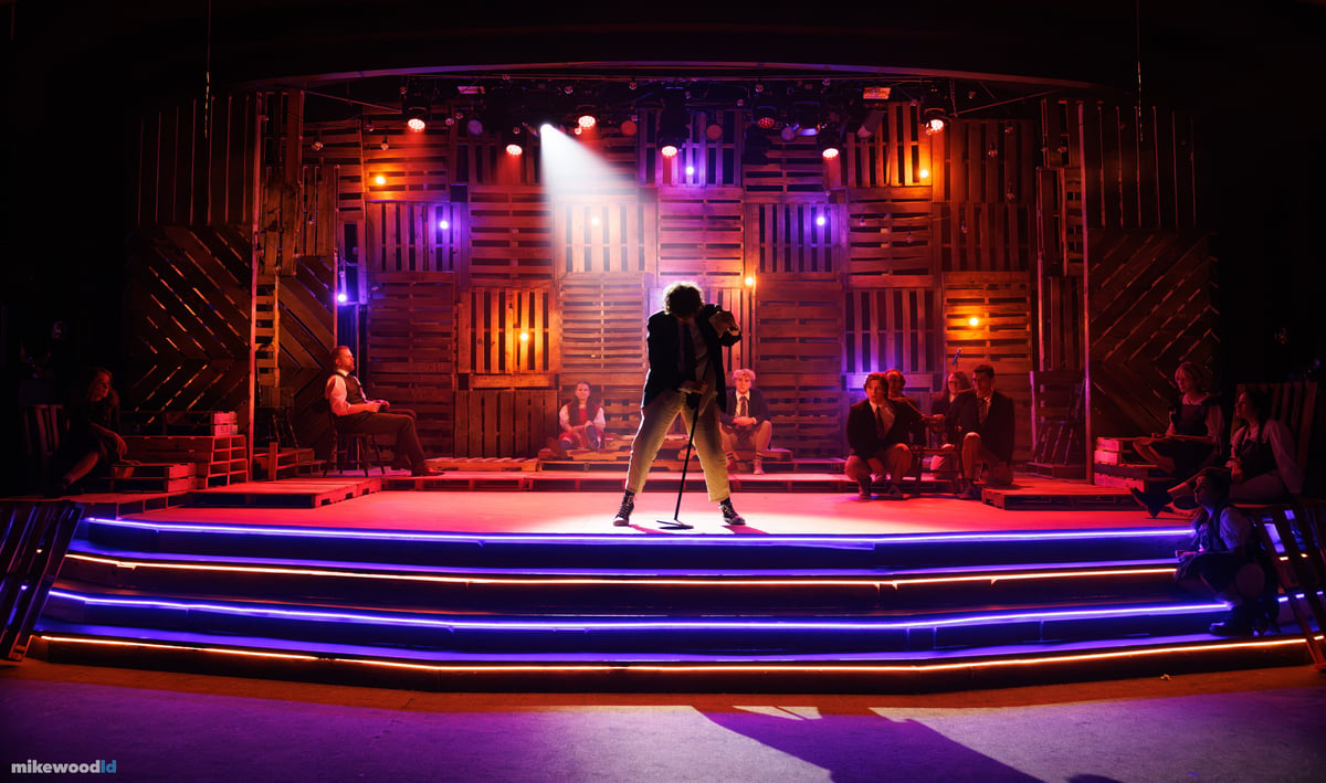 A lone young man stands silhouetted at a microphone under a single top special, the pallet wall behind him in warm amber and violet, blue LED striping outlining the steps.
