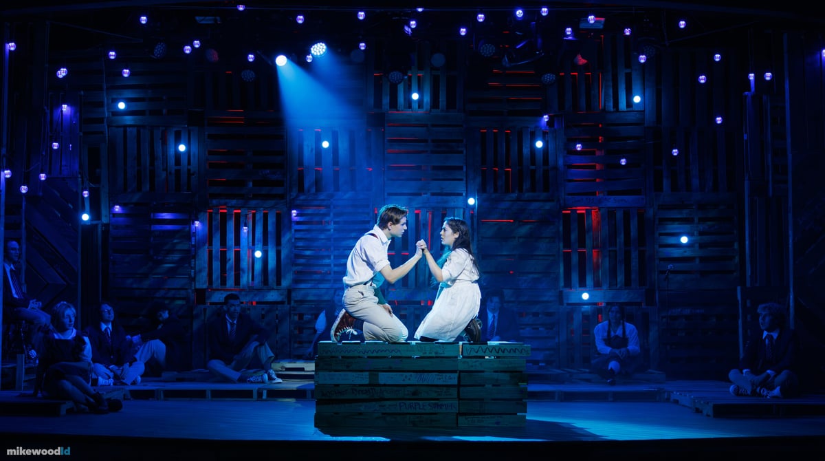 A young couple kneels facing each other, hands clasped, on a pallet crate in a tight blue special, the ensemble seated in the dark as red and blue practicals scatter across the wall.